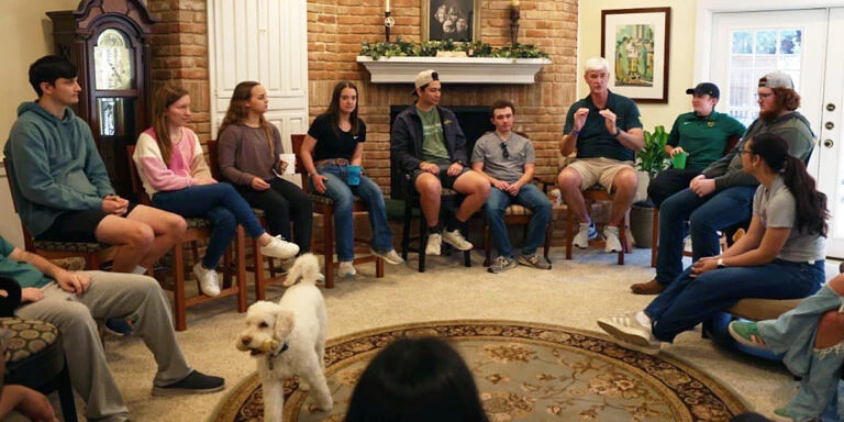 A group of people is gathered in a living room, sitting in a circle with a dog in the center.
