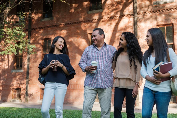 A family stands together looking at each other and smiling on grass outside a brick building, while holding coffee, book bags, and books.